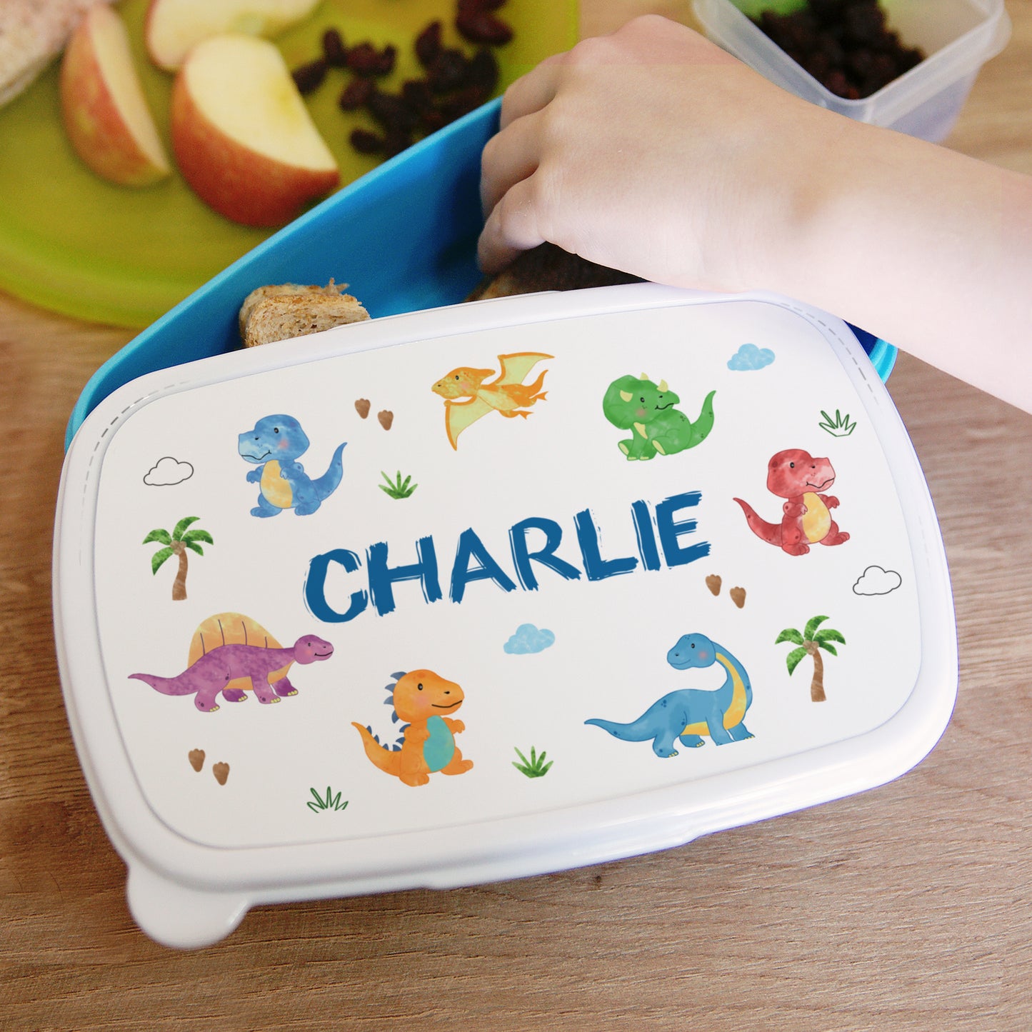 Children's lunchbox with dinosaur designs and the name 'CHARLIE' on a wooden table.