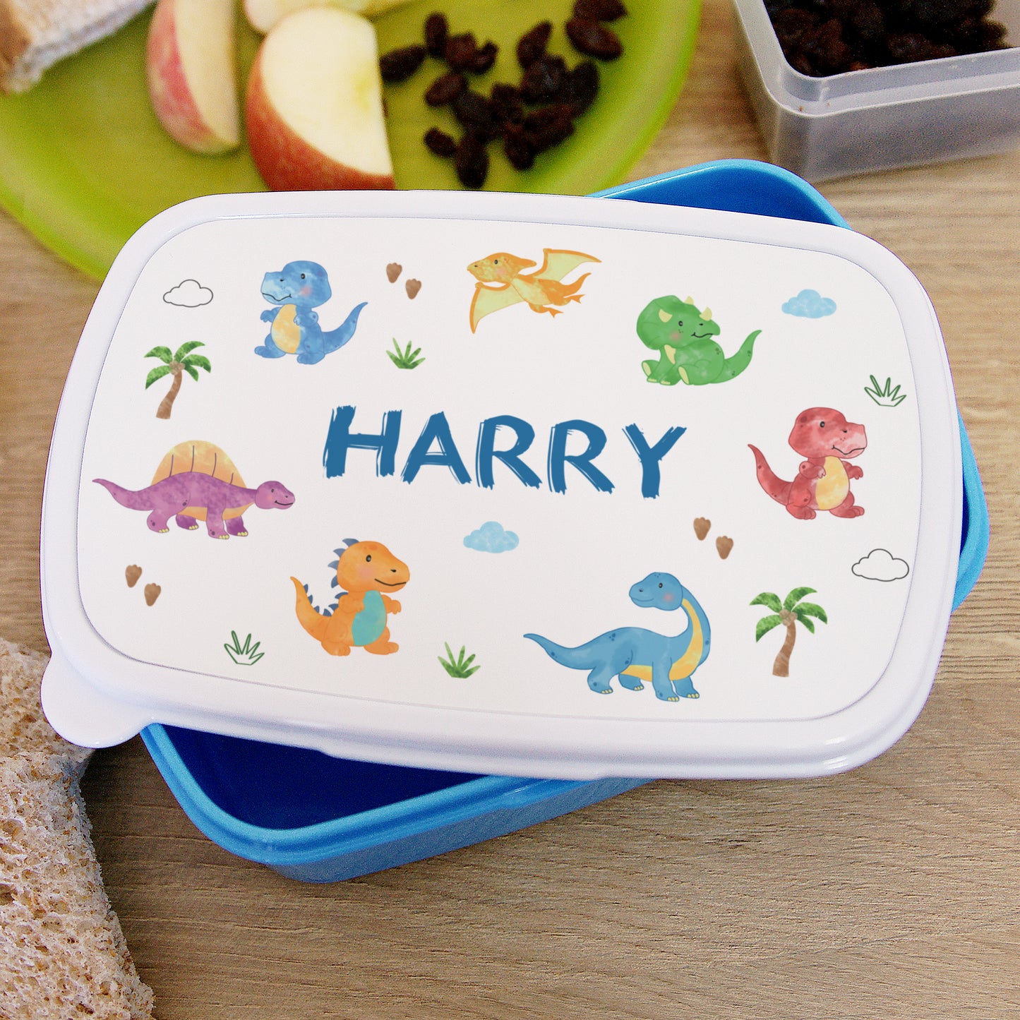 Children's lunchbox with dinosaur design and 'Harry' printed on it, placed on a wooden surface with snacks.