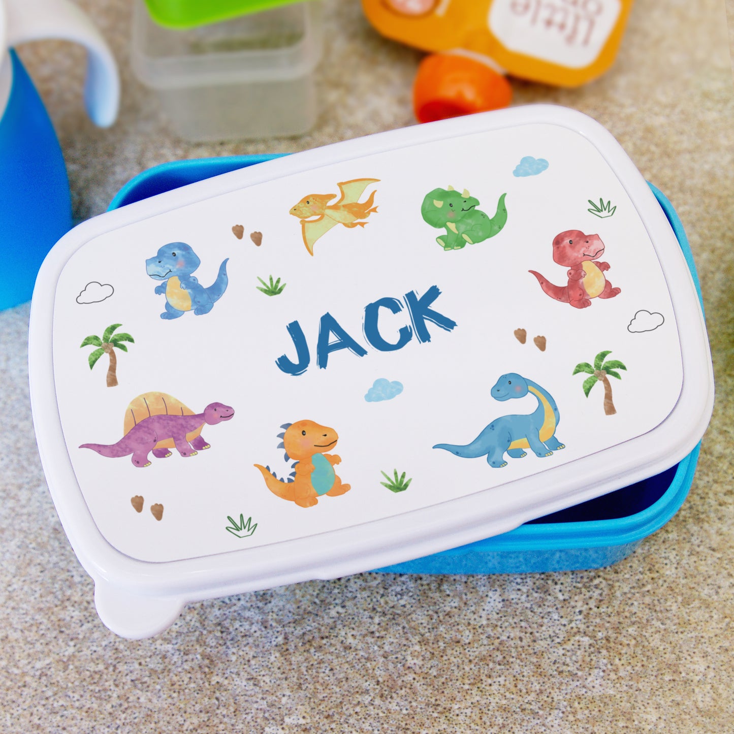Children's lunch box with dinosaur illustrations and the name 'Jack' on a sandy background