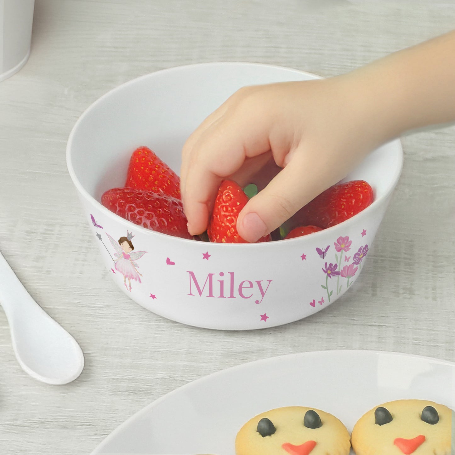 Child's hand reaching into a bowl of strawberries with 'Miley' printed on it, on a light wooden surface.