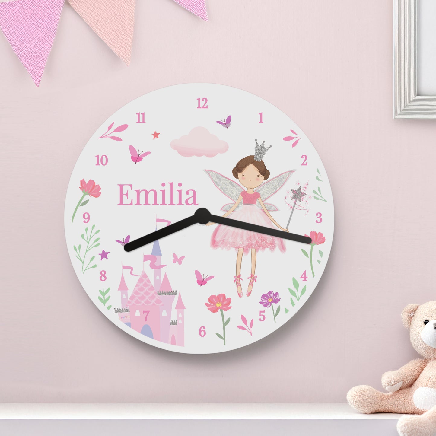 Children's wall clock with fairy design and 'Emilia' on a pink wall with a teddy bear.