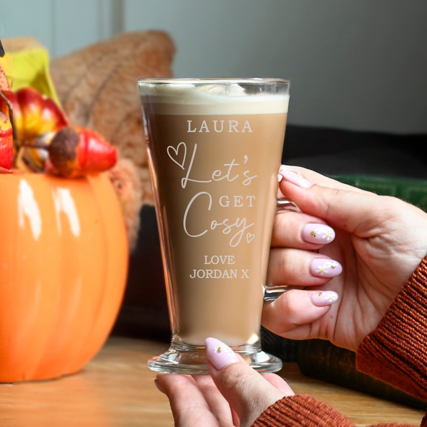 Person holding a glass of coffee with personalised text, next to a pumpkin and autumn decorations.