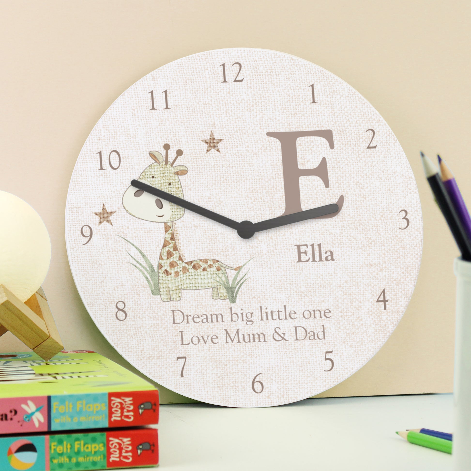 Personalised clock shown on a shelf alongside books and pencils. Text reads Ella Dream big little one Love Mum & Dad
