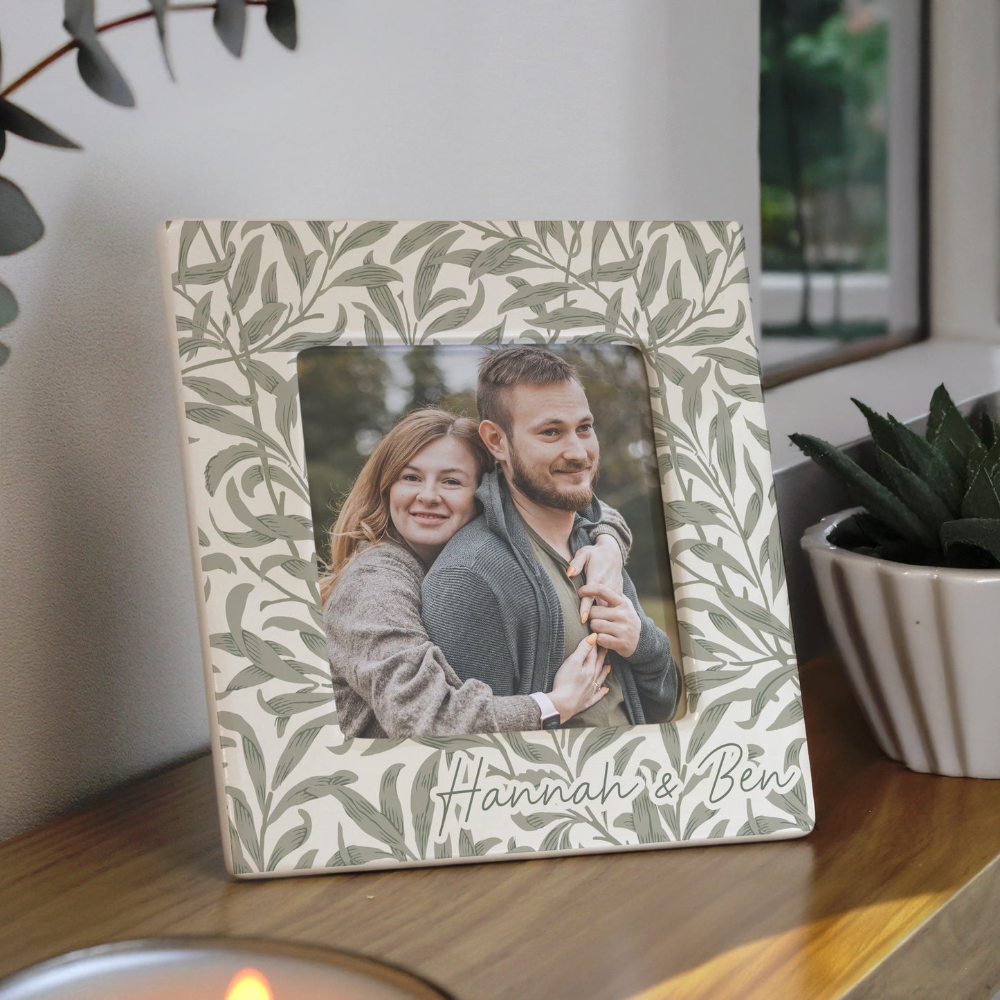Decorative photo frame with a couple's photo and 'Hannah & Ben' text, placed on a surface with plants in the background.