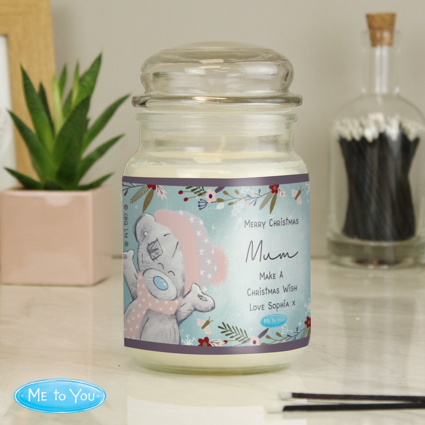 Candle in a glass jar with a decorative label featuring an cute bear design, placed on a surface with a plant and another jar in the background.