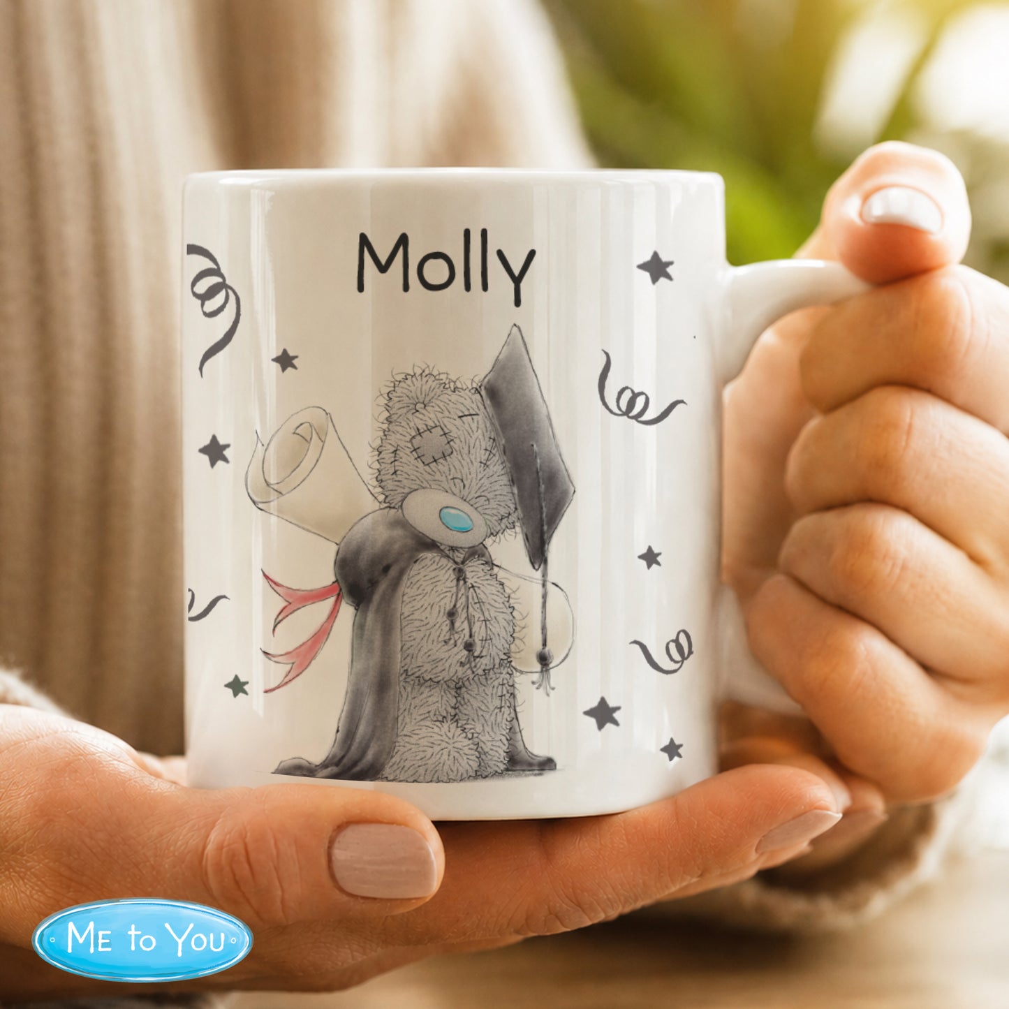Mug with a teddy bear design and 'Molly' text, held by hands with a blurred background.