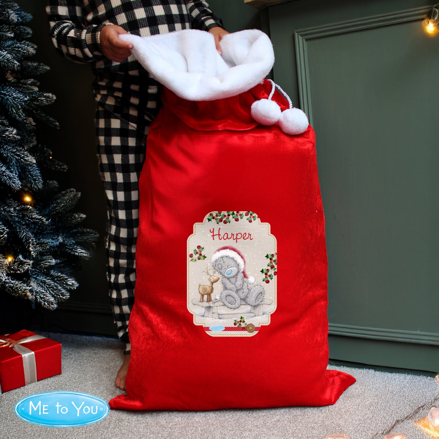Red Christmas sack with 'Harper' and a teddy bear design, held by a person in checkered pajama pants.