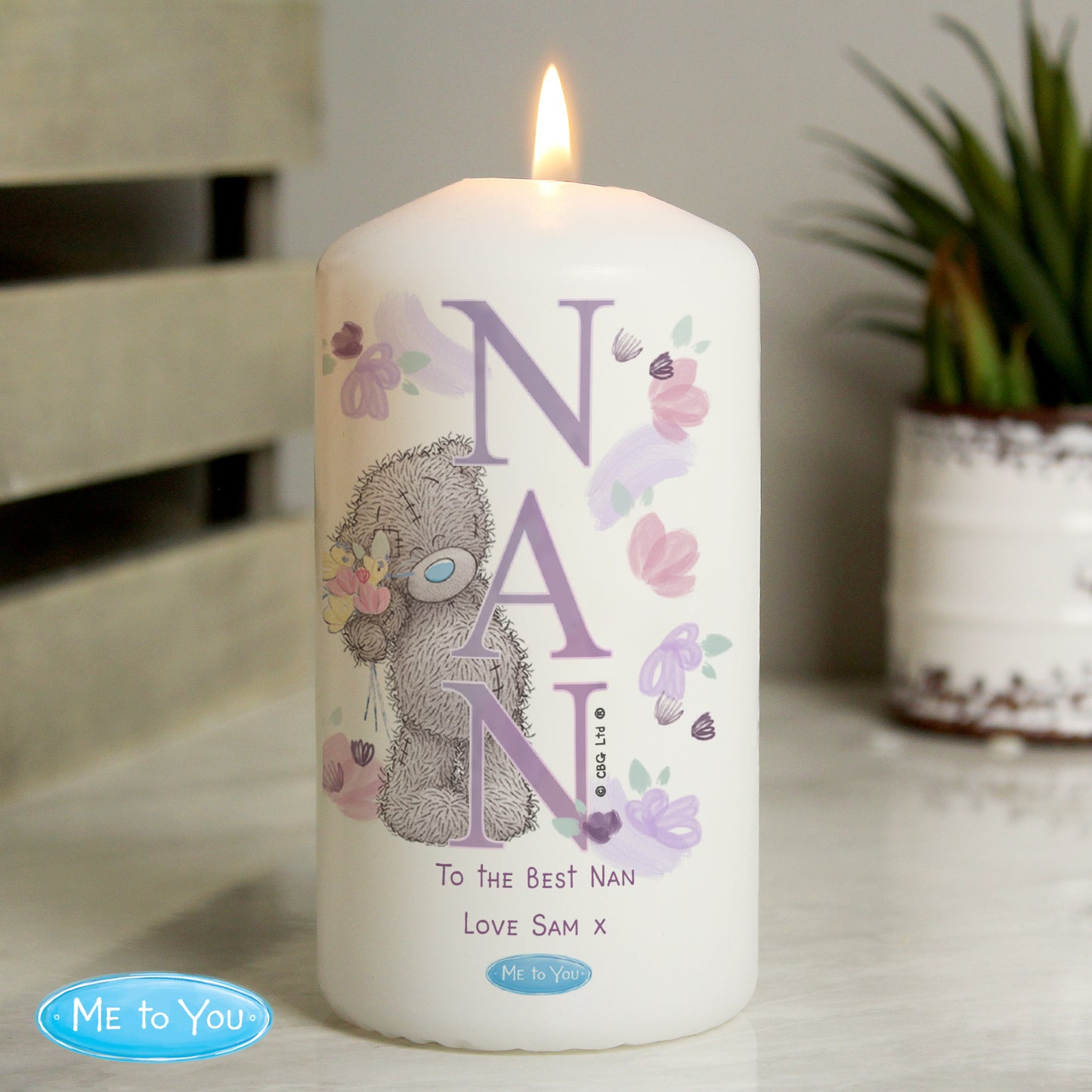Candle with 'NAN' and a bear design, featuring the Me to You brand, on a surface with a plant in the background.