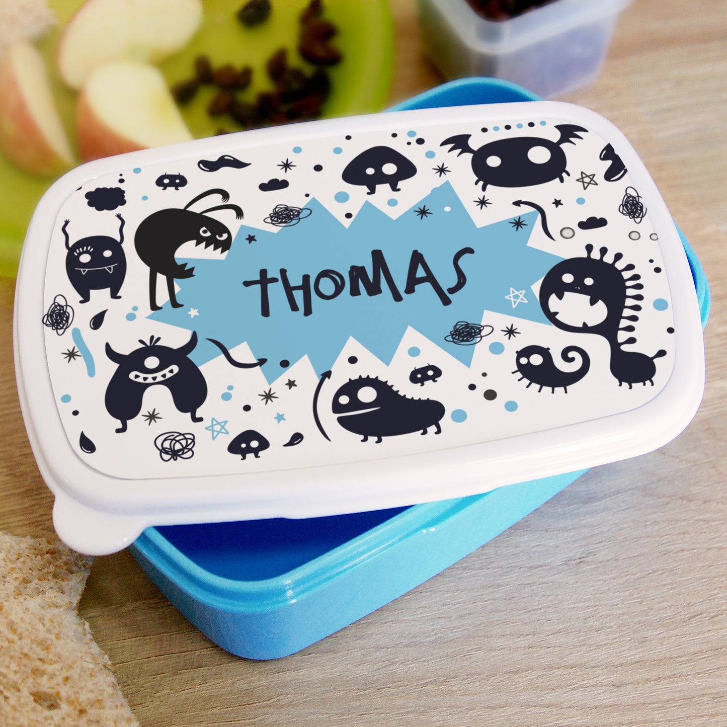 Children's lunch box with monster design and 'THOMAS' branding on a wooden surface.