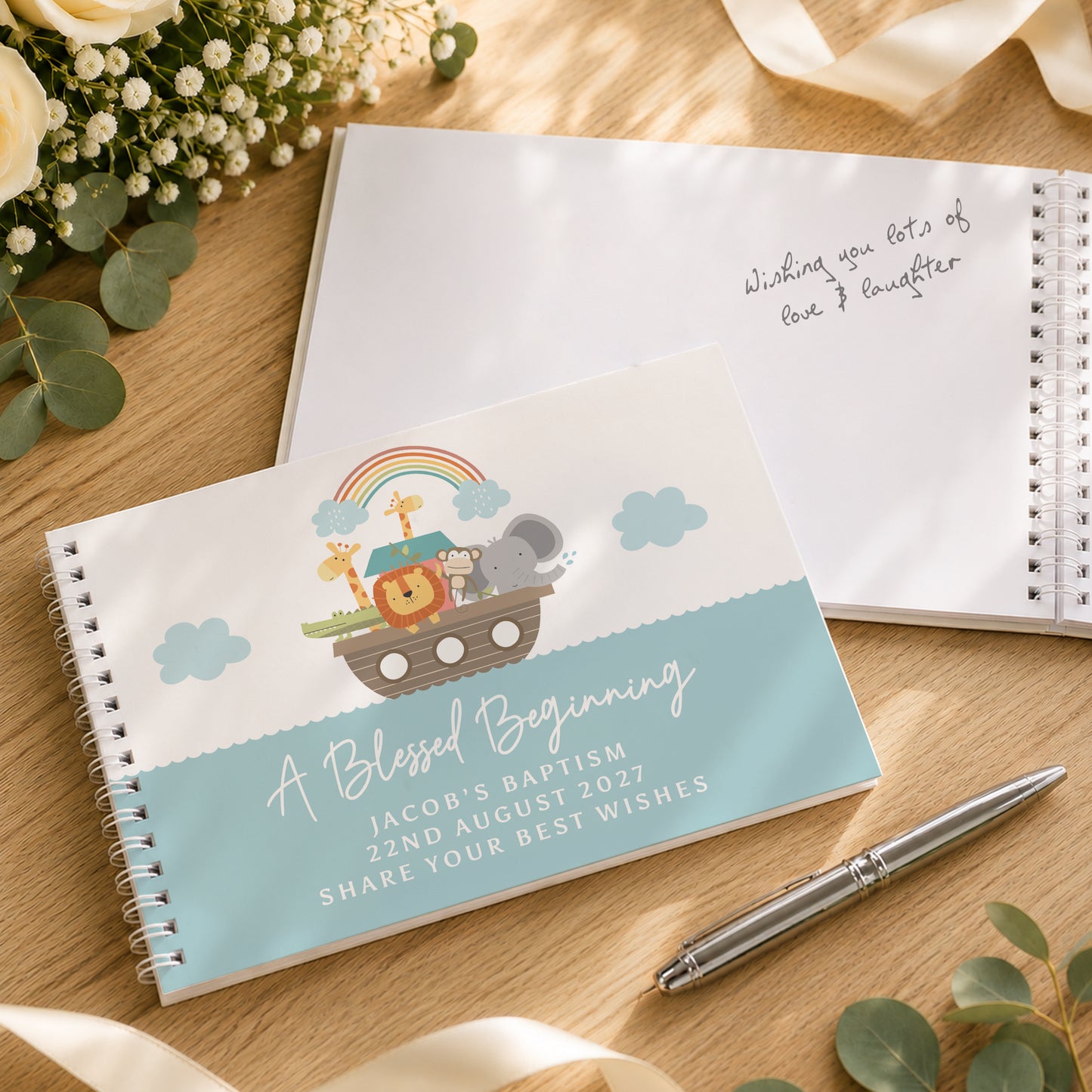 A personalised baby baptism keepsake guest book with a light blue and white cover featuring a Noah's Ark cartoon illustration, shown on a wooden table next to a matching open notebook with handwritten wishes, a silver pen, and eucalyptus leaves.