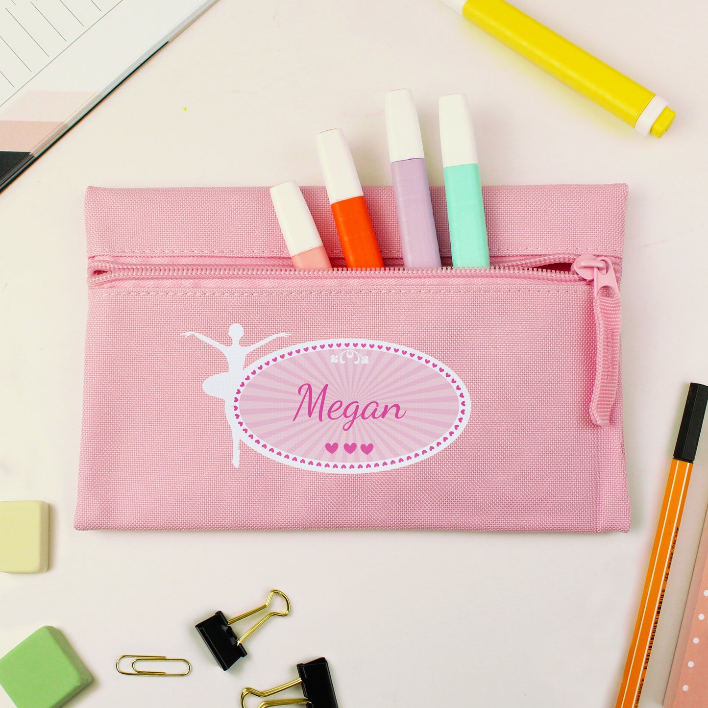Pink pencil case with name 'Megan' and ballet dancer design, surrounded by stationery items on a light background.