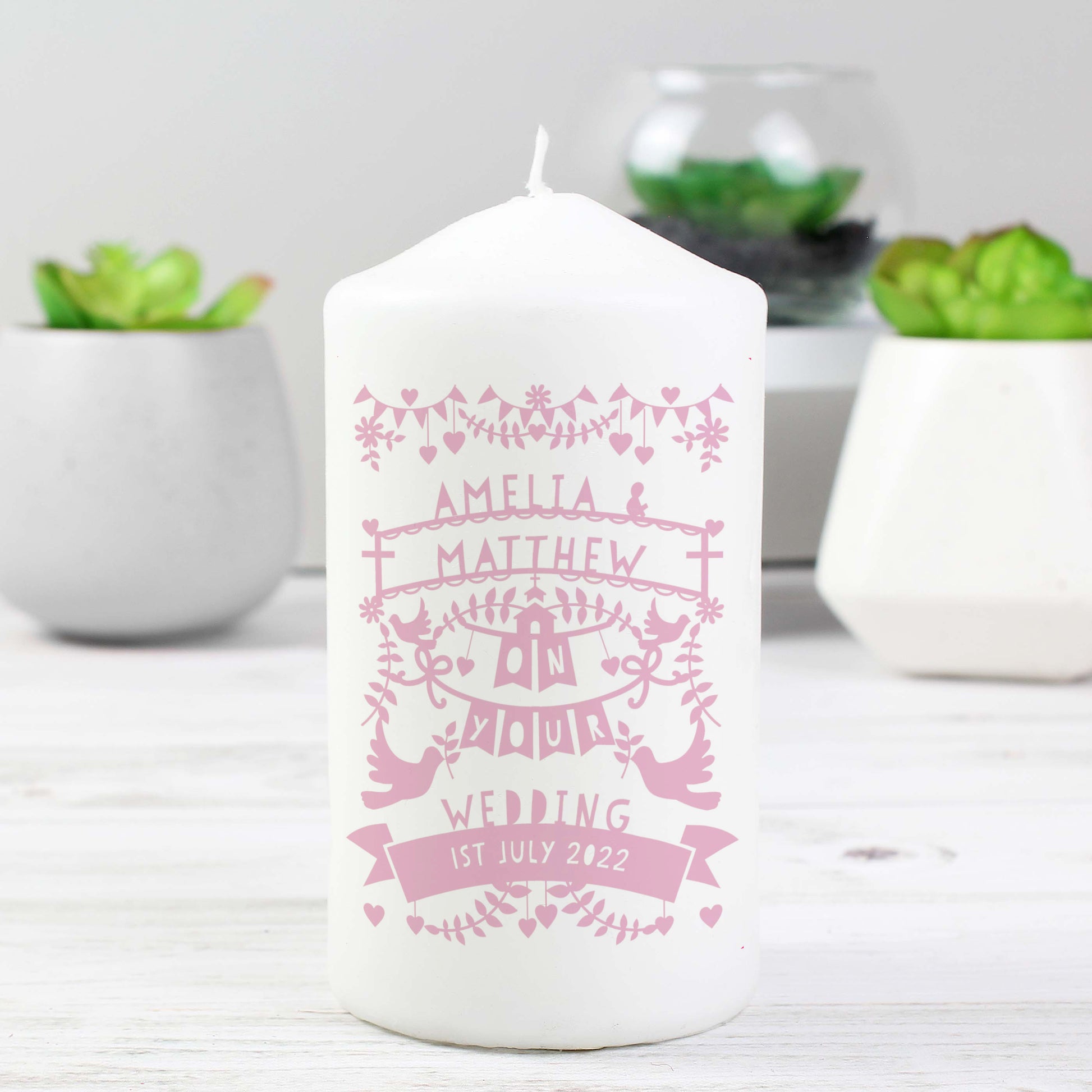 White candle with pink personalised text on a white surface with plants in the background