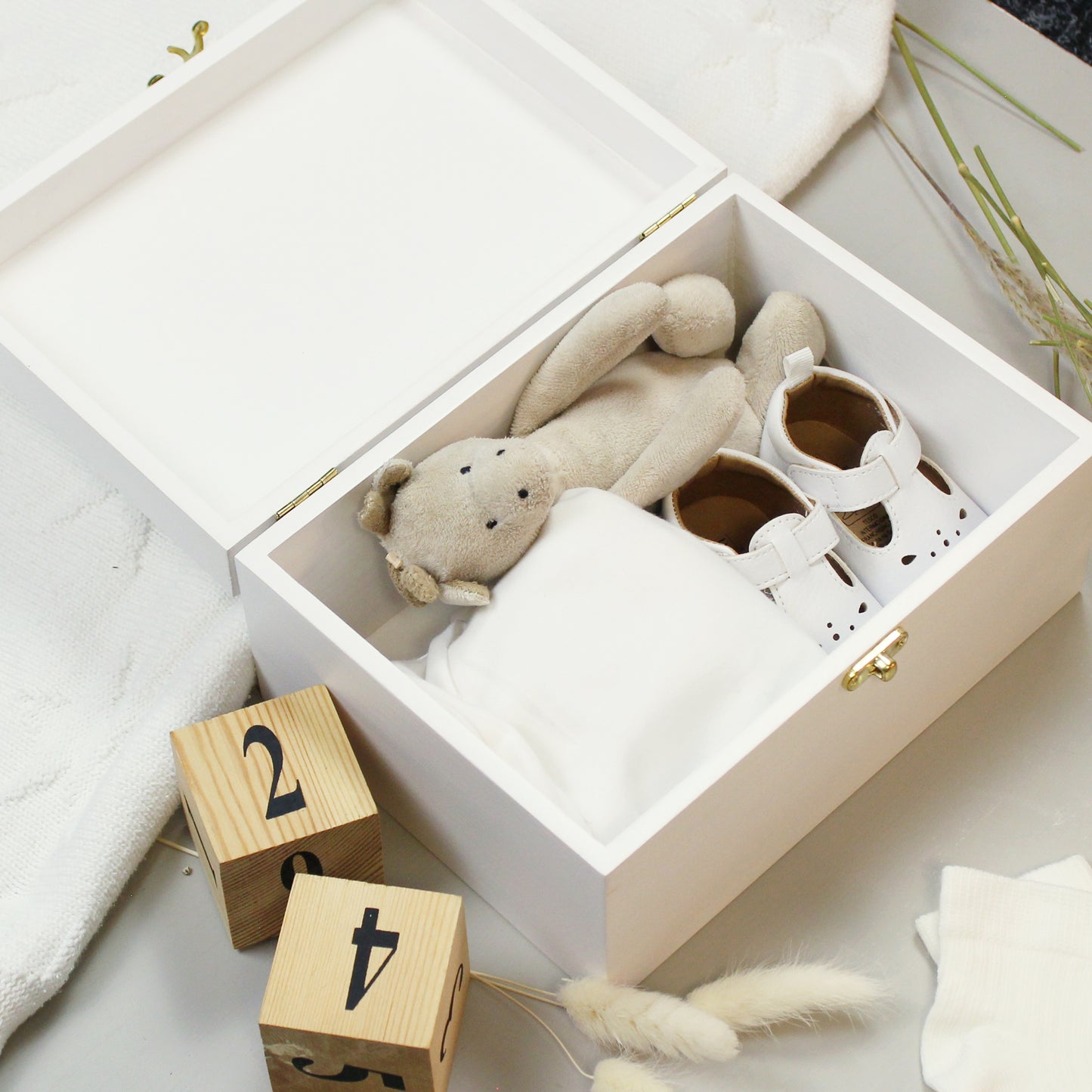 White box with baby items including a teddy bear, booties, and blocks on a light surface.
