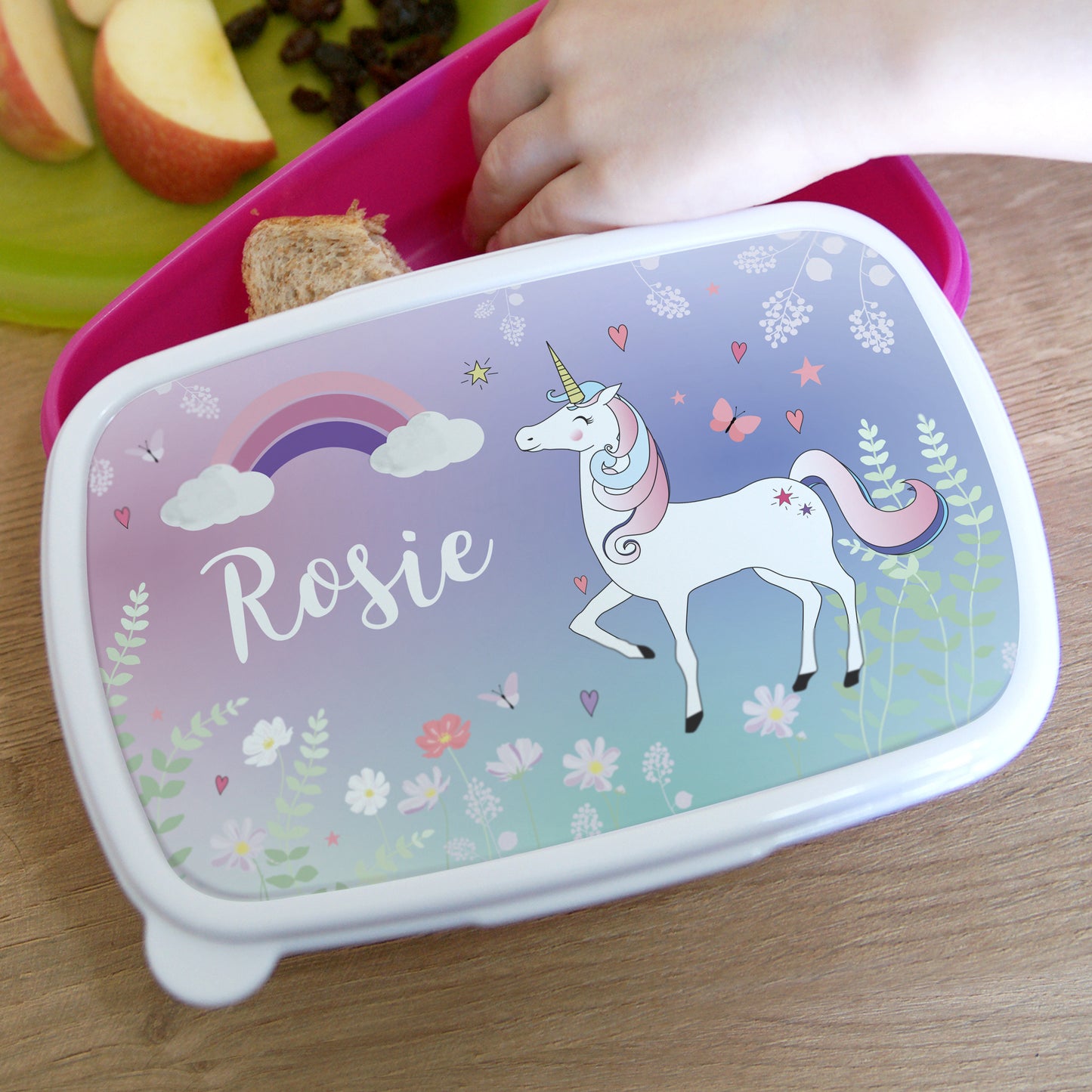 Lunch box with unicorn design and 'Rosie' text, placed on a wooden surface with a snack.