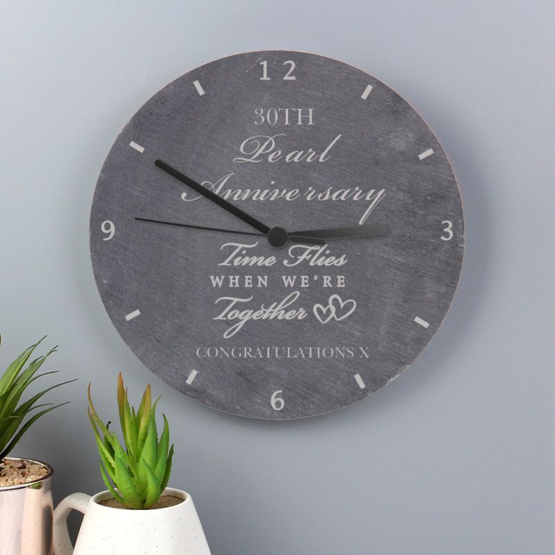 Wall clock with personalised message on a grey wall with plants below.