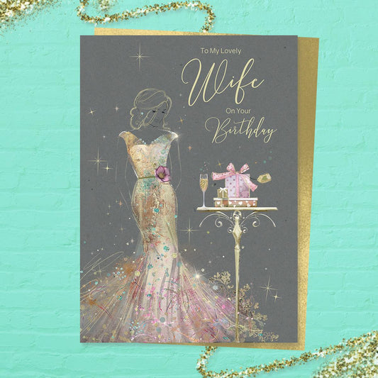 To My Lovely Wife On Your Birthday' card featuring a beautiful lady in a long gold dress with champagne and gifts. With added sparkle and gold foil detail. Colour image inside with heartfelt verse. Complete with gold colour envelope