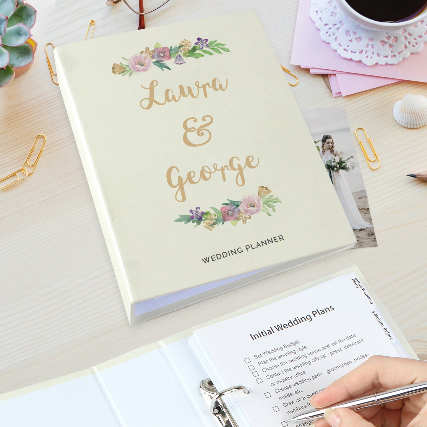 Wedding planner book with floral design and 'Laura & George' text, on a desk with stationery items.