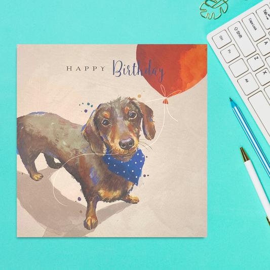 Square card with sausage dog in neck scarf holding a red balloon