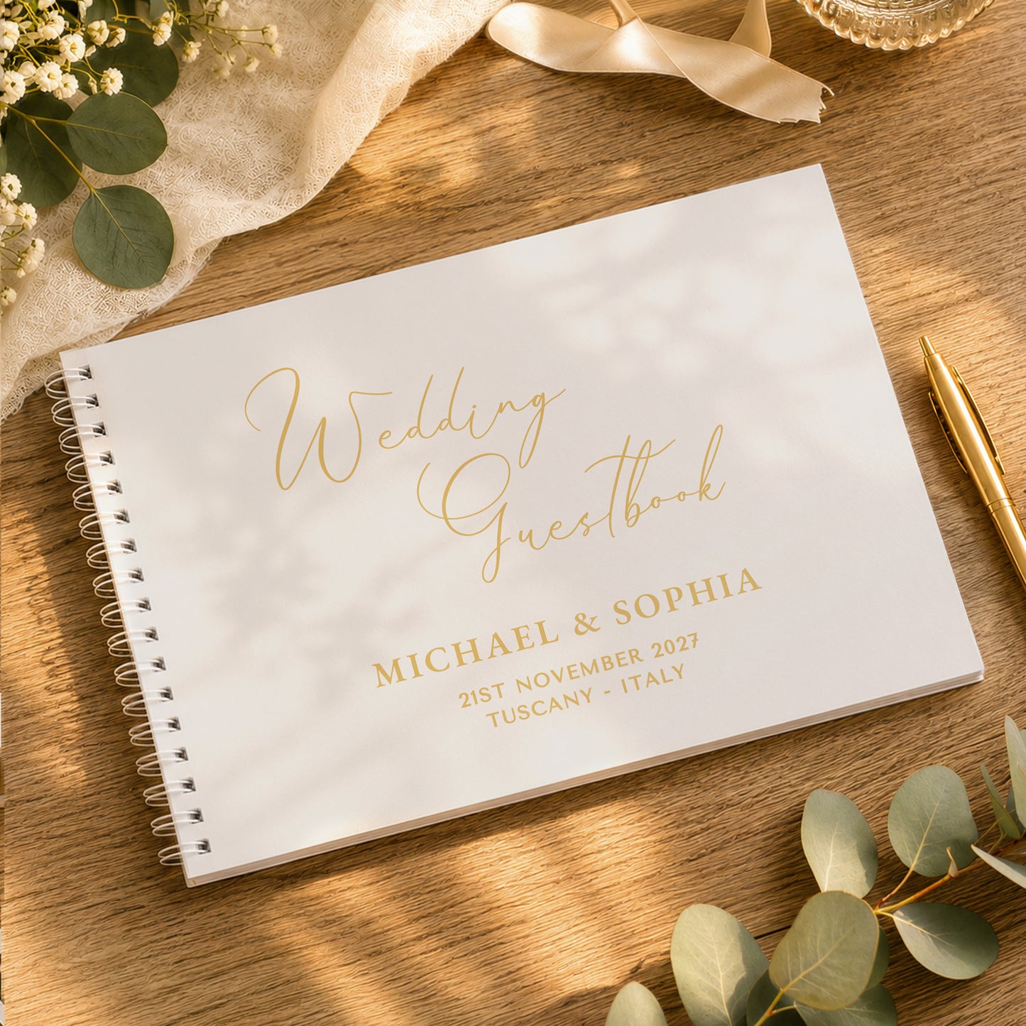 A personalised white spiral-bound wedding guestbook with elegant gold foil lettering, styled on a wooden tabletop with eucalyptus leaves, a gold pen, and soft decorative props.