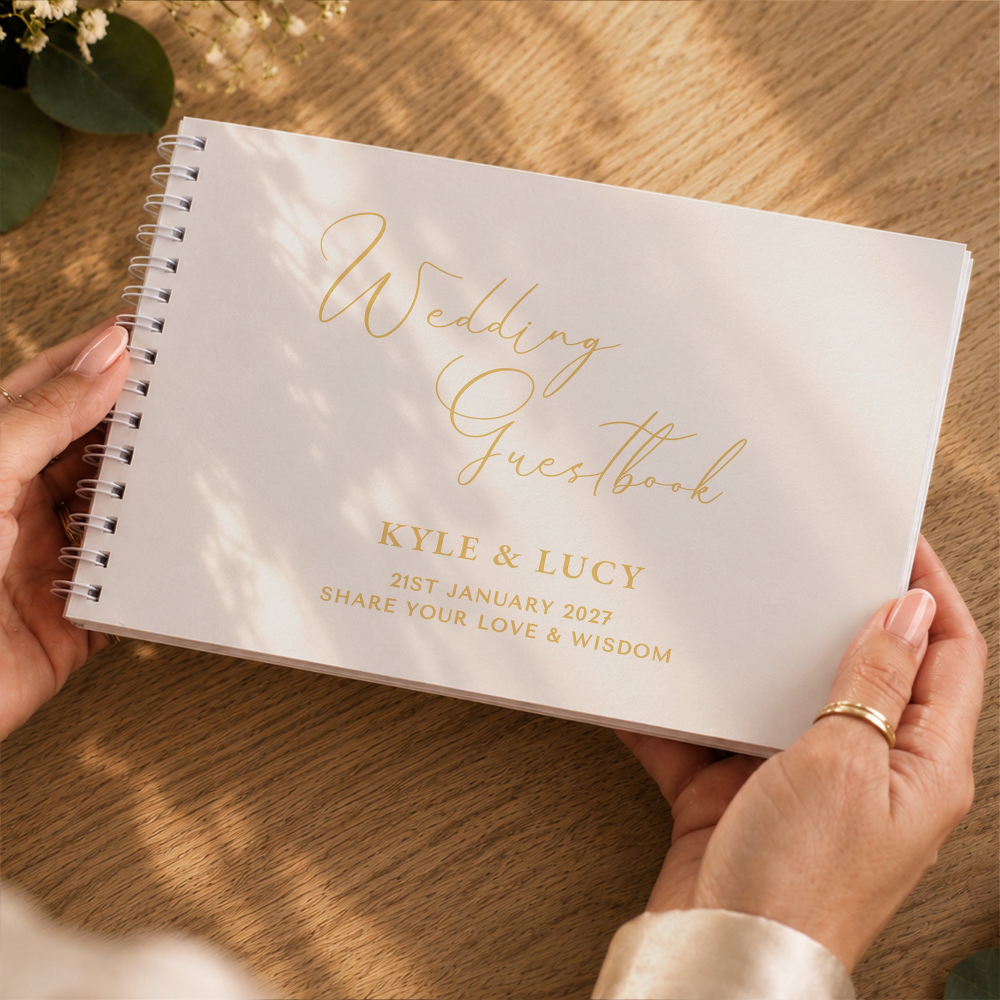 A personalised white spiral-bound wedding guestbook held by two hands over a wooden table. The guestbook features elegant gold foil script reading 'Wedding Guestbook' and gold foil text reading 'Kyle & Lucy 21st January 2027 Share Your Love & Wisdom'.