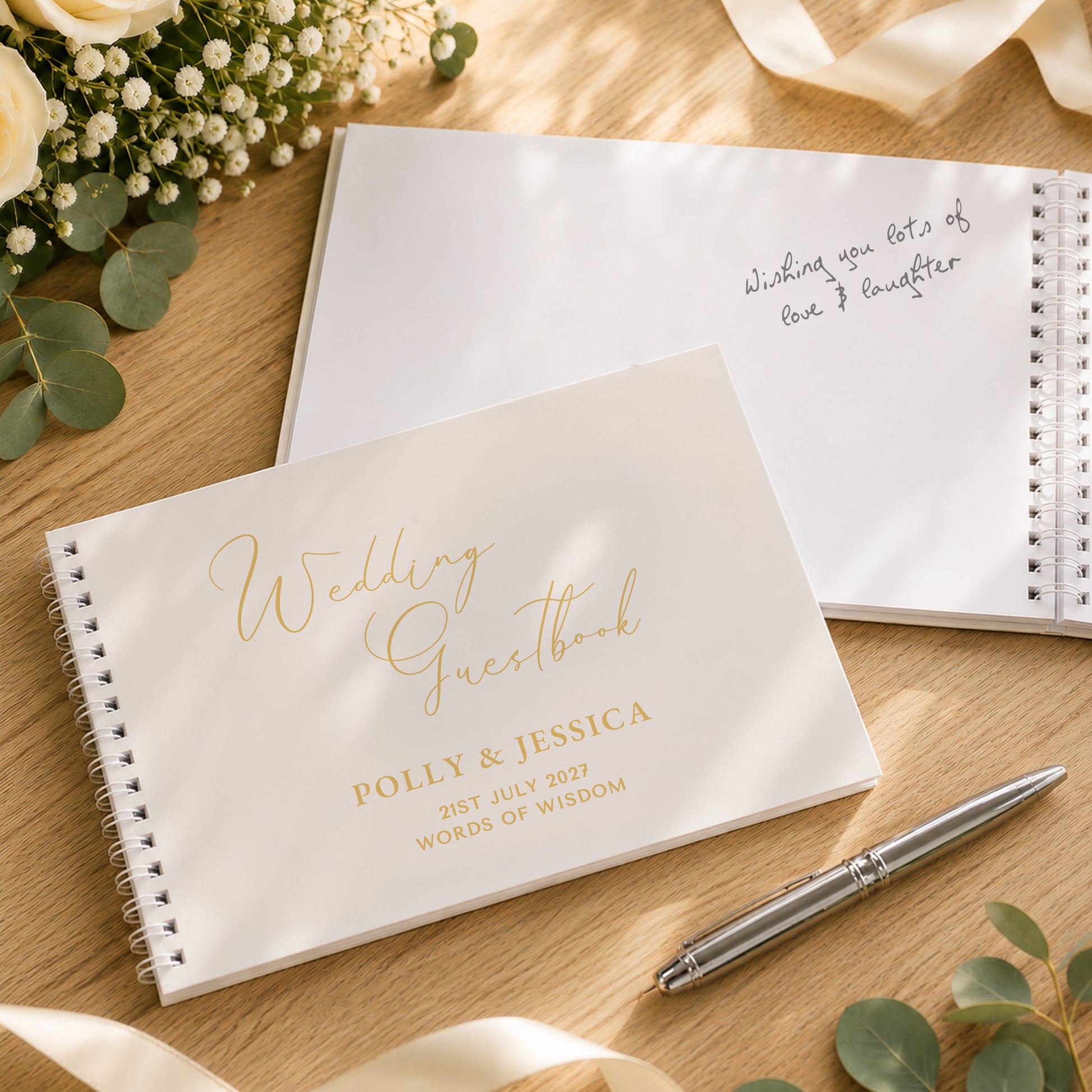A styled flat-lay of two wedding guestbooks on a wooden surface. The foreground guestbook is white with elegant gold foil script reading 'Wedding Guestbook' and personalized names/date text.