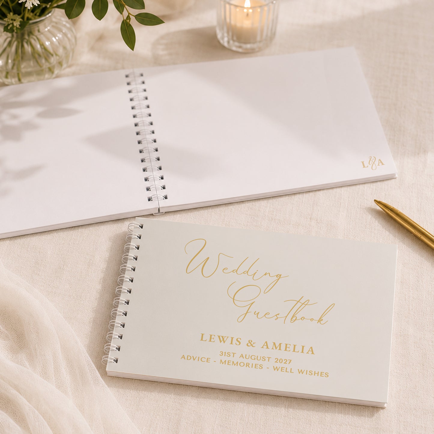 A personalised wedding guestbook notebook lies open on a cream fabric surface, with another closed copy in front showing gold foil text that reads 'Wedding Guestbook' and names/date details. A gold pen, candle, and flowers are softly styled around it.
