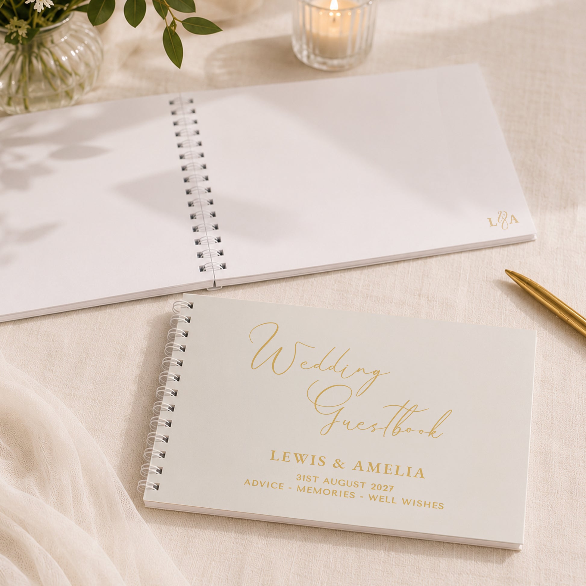 A personalised wedding guestbook notebook lies open on a cream fabric surface, with another closed copy in front showing gold foil text that reads 'Wedding Guestbook' and names/date details. A gold pen, candle, and flowers are softly styled around it.
