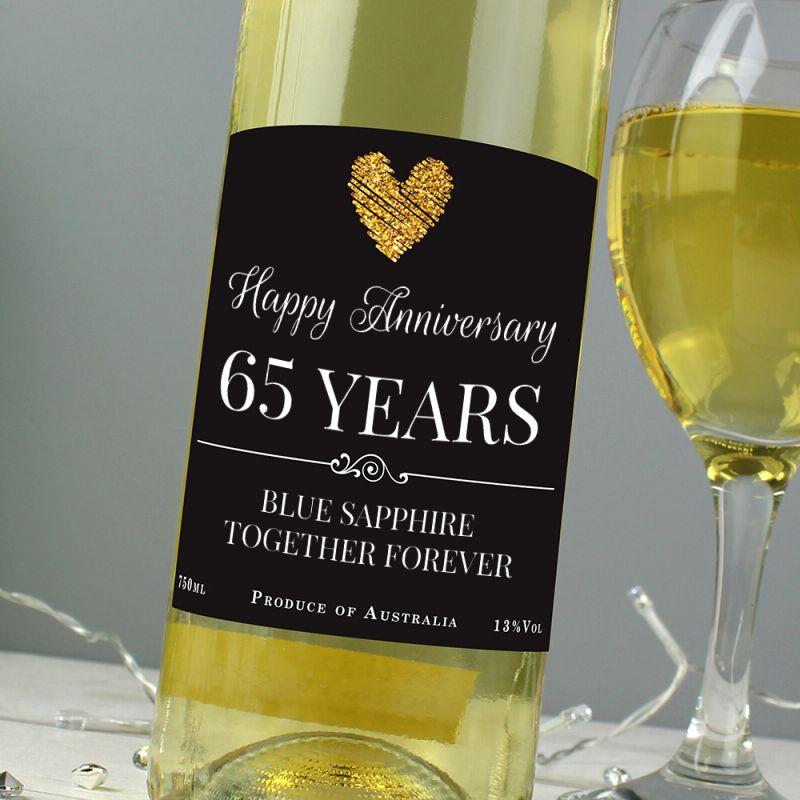 Bottle of white wine with a 'Happy Anniversary 65 Years' label next to a glass of wine on a gray background.
