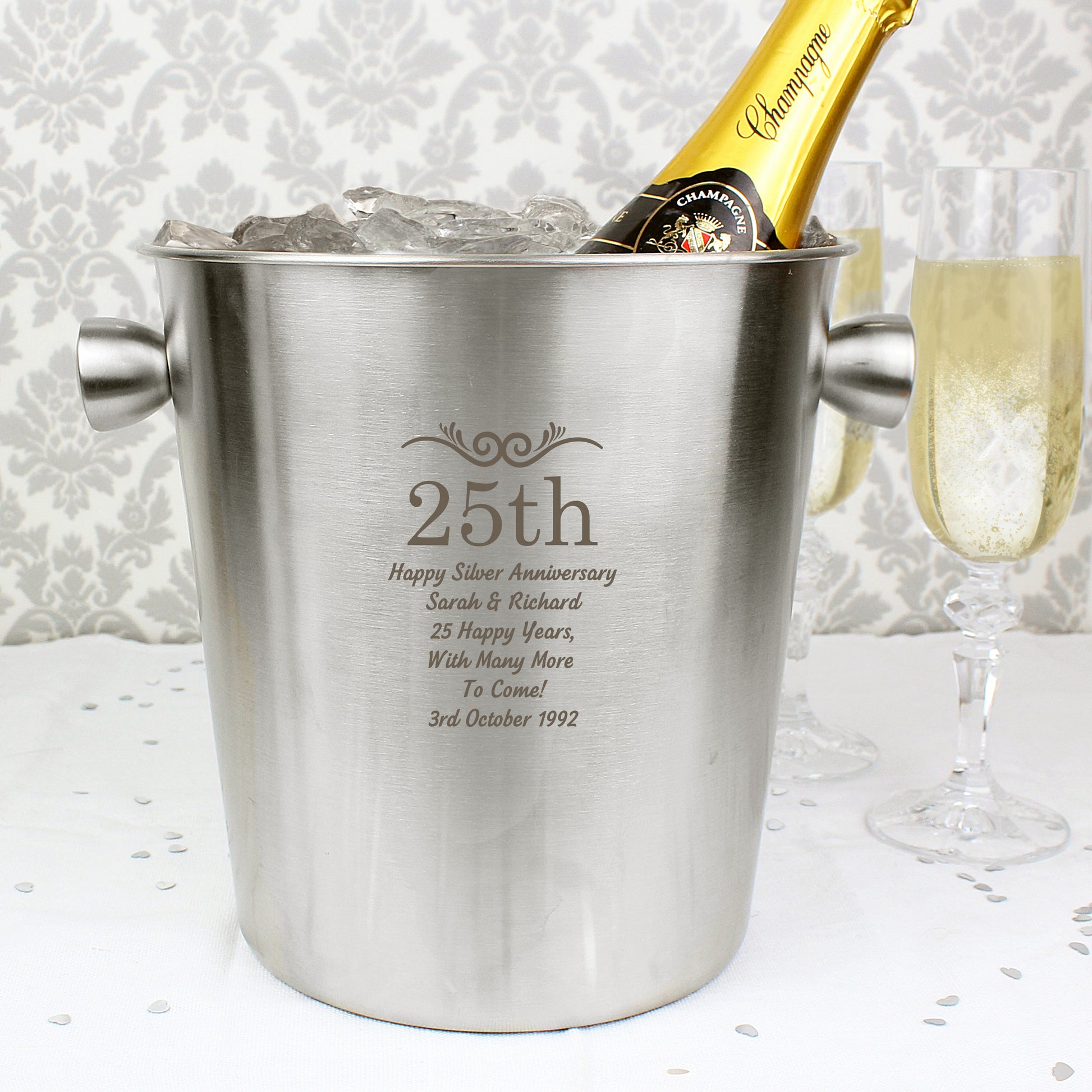 A silver stainless steel ice bucket with personalized text '25th' and a message celebrating an anniversary. It is placed on a table with a glass of champagne next to it.