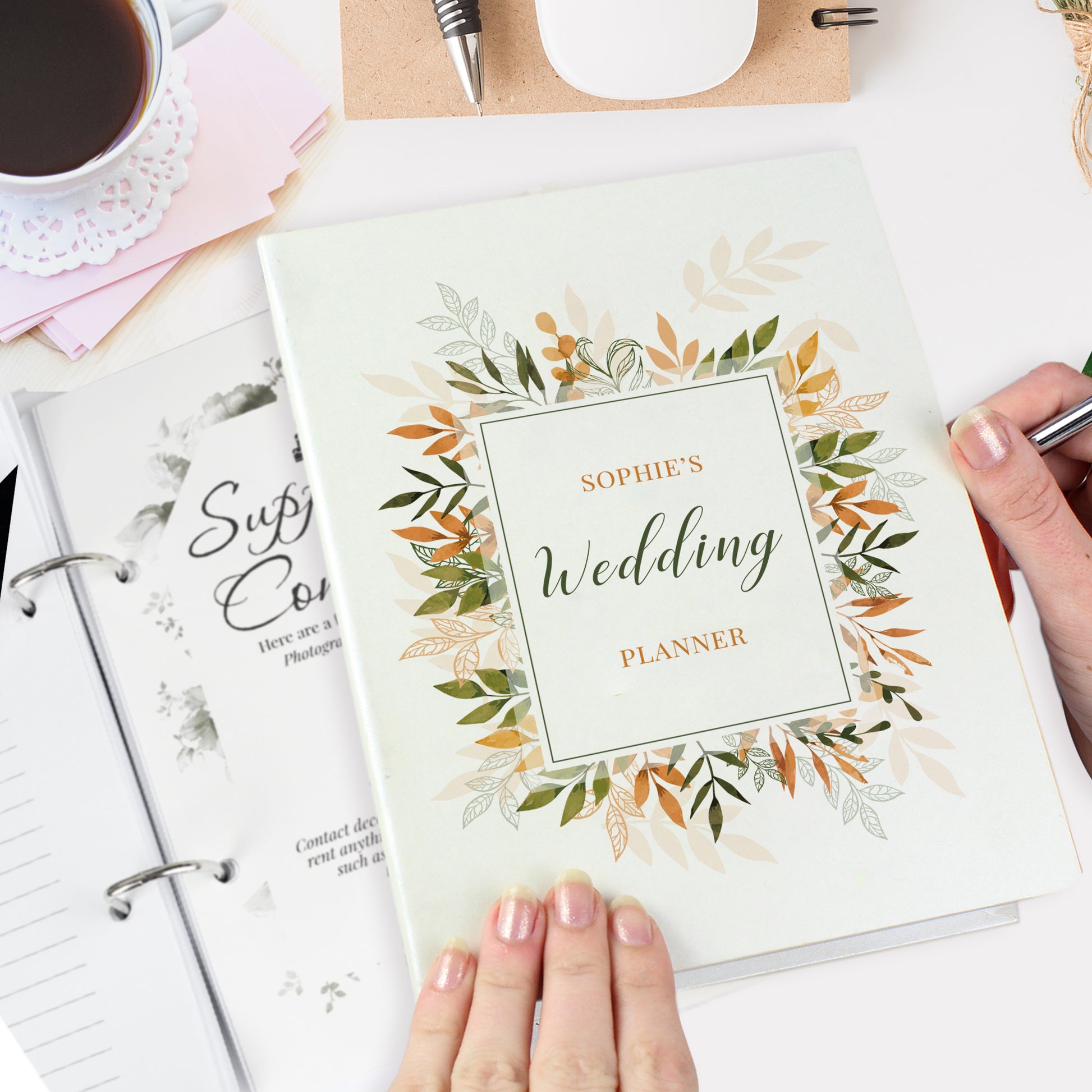 Wedding planner book with floral design held by a hand, surrounded by stationery items.