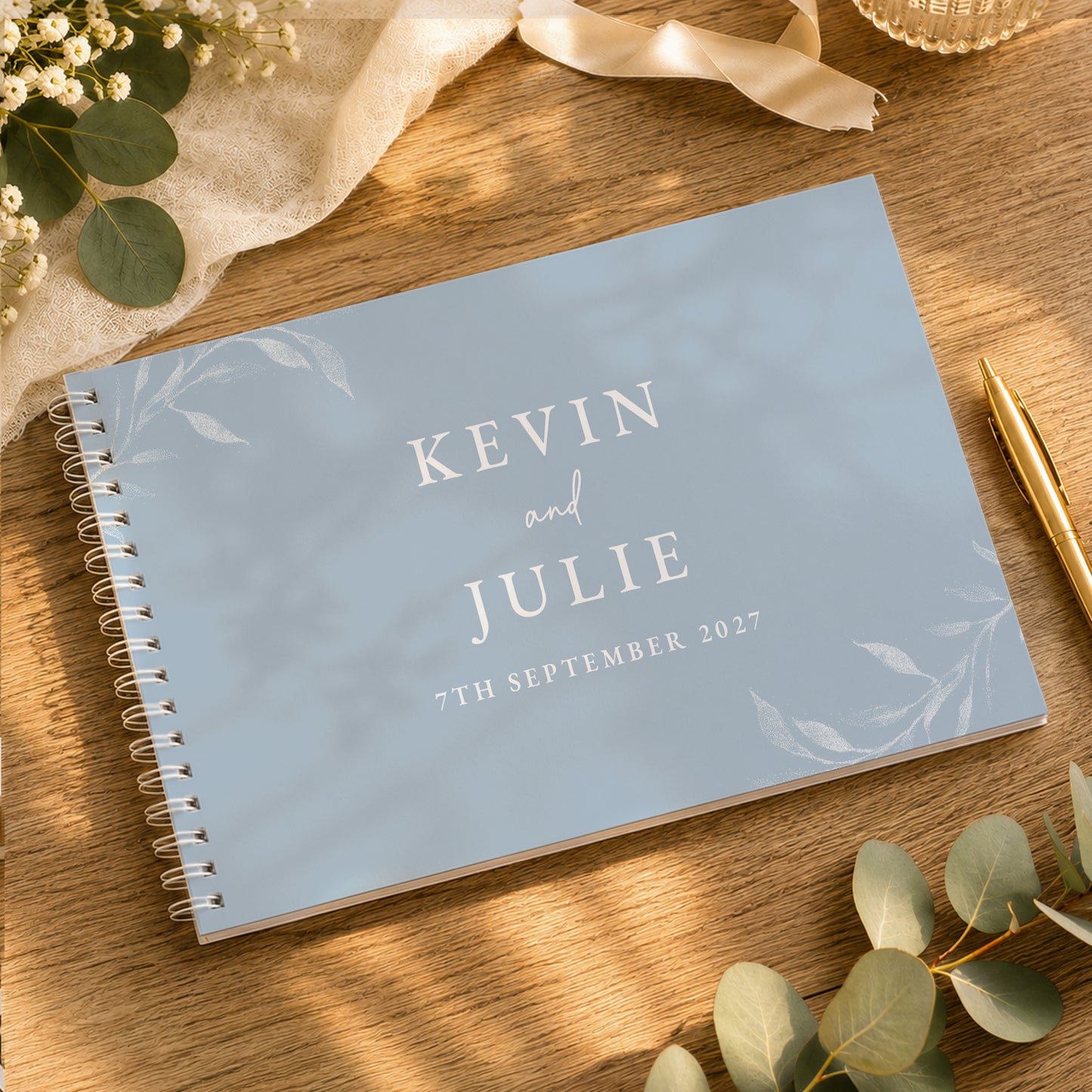 A personalised spiral-bound guest book with a light blue cover and white text reading 'KEVIN and JULIE 7TH SEPTEMBER 2027', styled on a wooden table with eucalyptus leaves, a gold pen, and cream fabric.