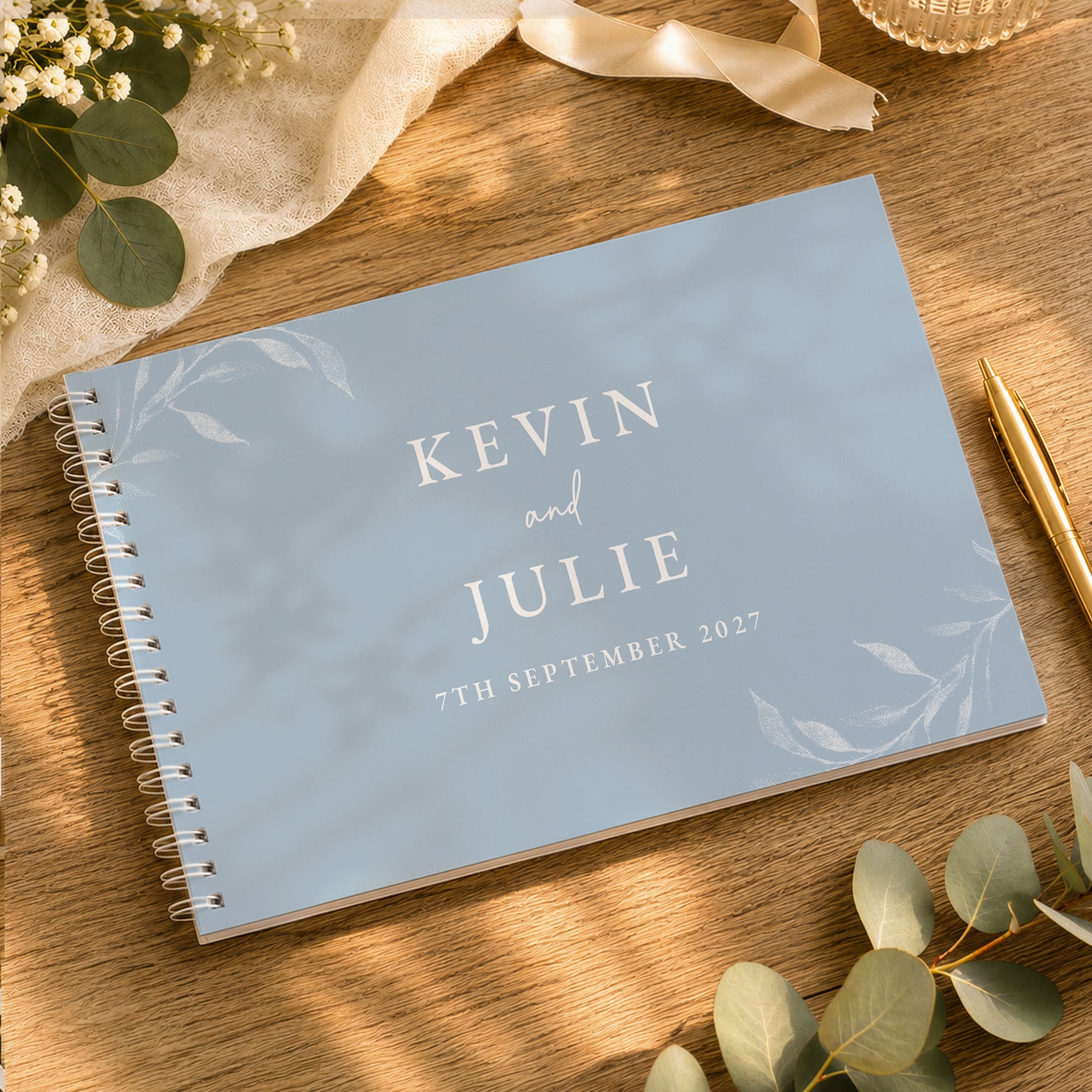 A personalised spiral-bound guest book with a light blue cover and white text reading 'KEVIN and JULIE 7TH SEPTEMBER 2027', styled on a wooden table with eucalyptus leaves, a gold pen, and cream fabric.