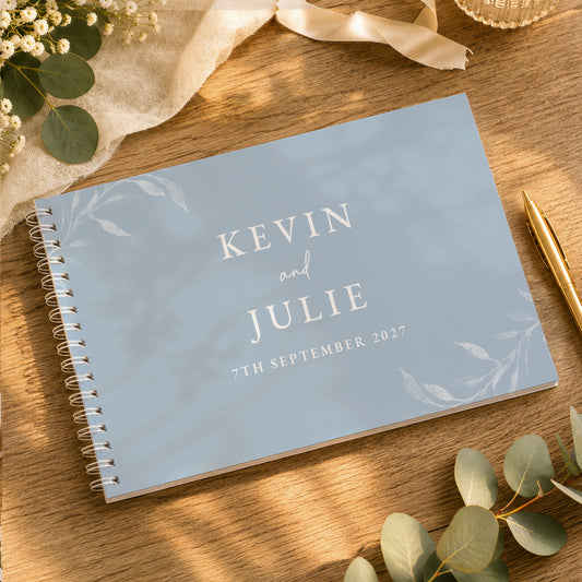 A personalised spiral-bound guest book with a light blue cover and white text reading 'KEVIN and JULIE 7TH SEPTEMBER 2027', styled on a wooden table with eucalyptus leaves, a gold pen, and cream fabric.