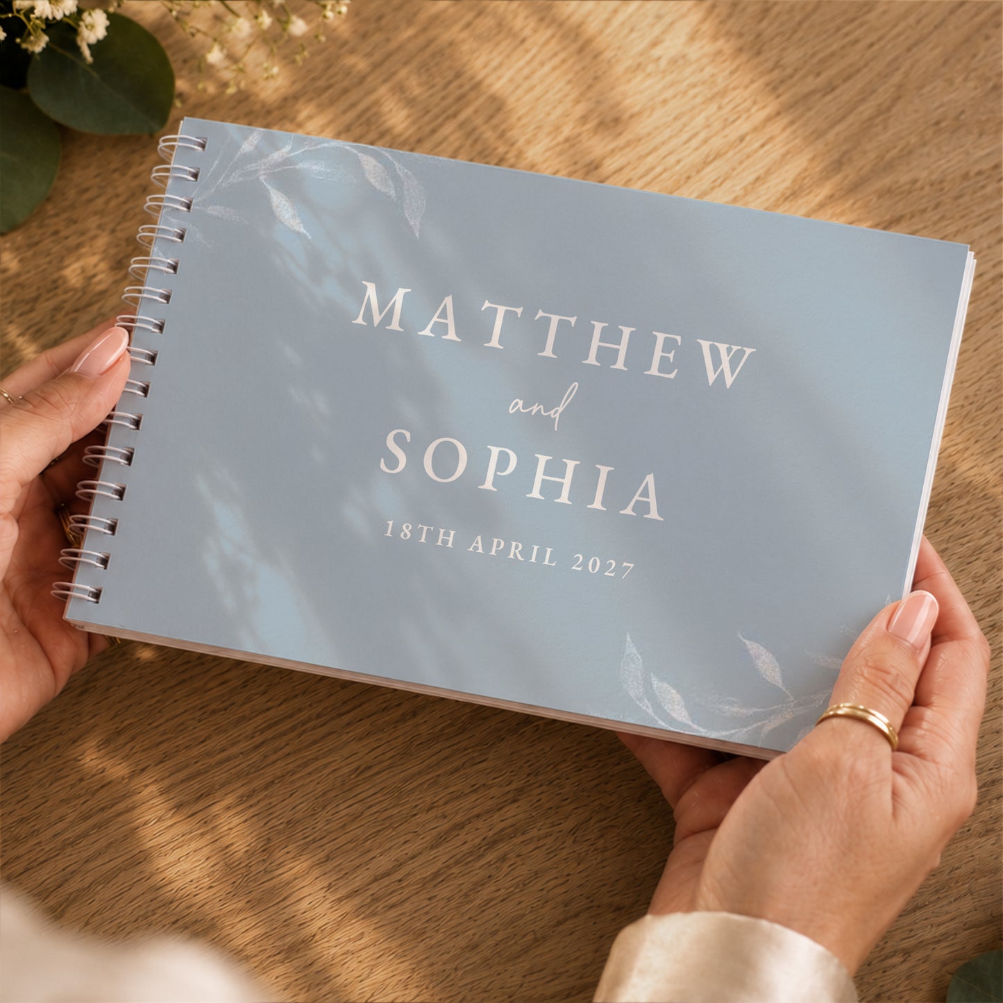 A personalised spiral-bound notebook held by two hands over a wooden table. The notebook cover is a dusty light blue with elegant white script reading 'MATTHEW and SOPHIA' and the date '18TH APRIL 2027'.
