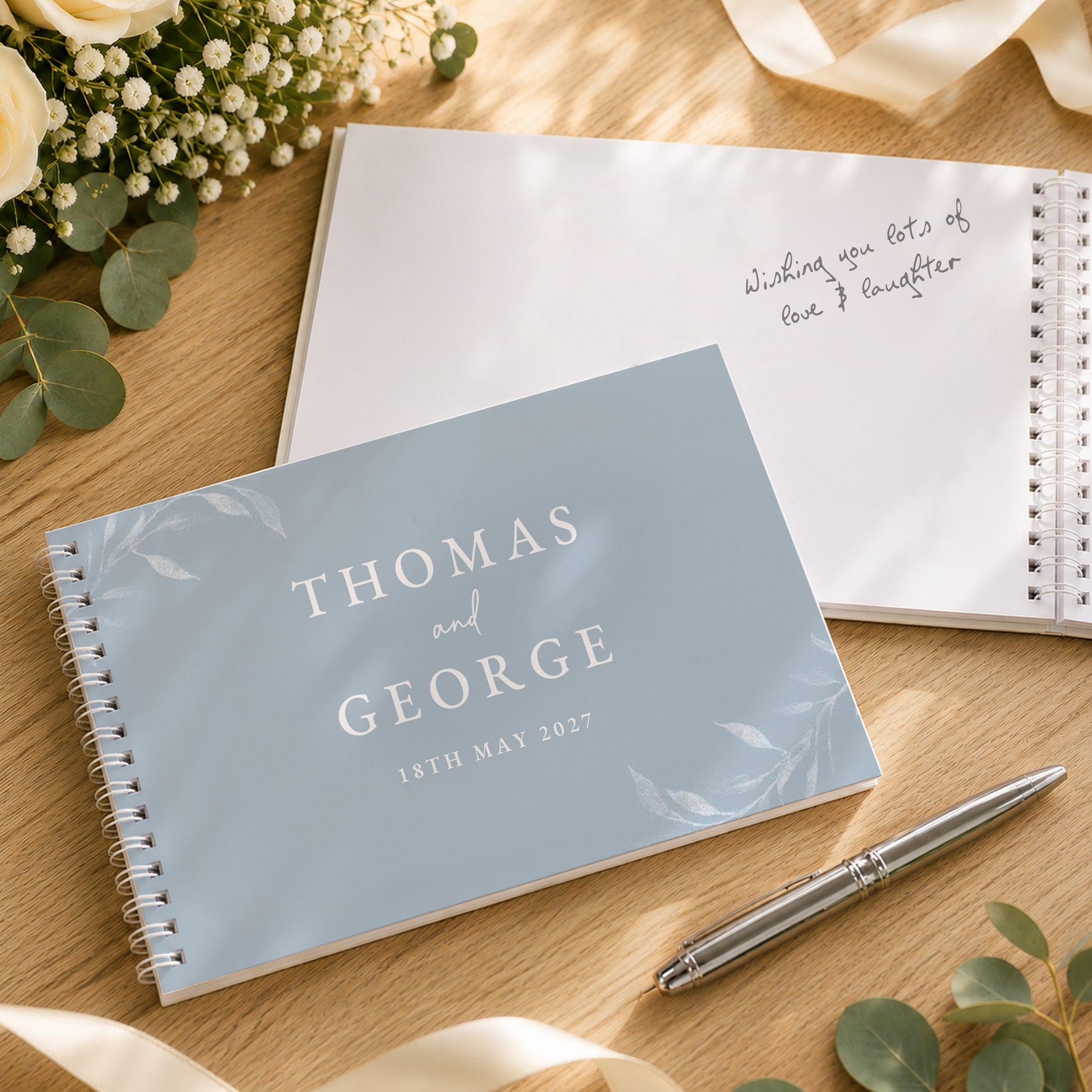 A personalised spiral-bound notebook with a pale blue cover reading 'THOMAS and GEORGE 18TH MAY 2027' is placed on a wooden surface, alongside an open white notebook with handwritten text and a silver pen. Floral and ribbon props are visible around it.