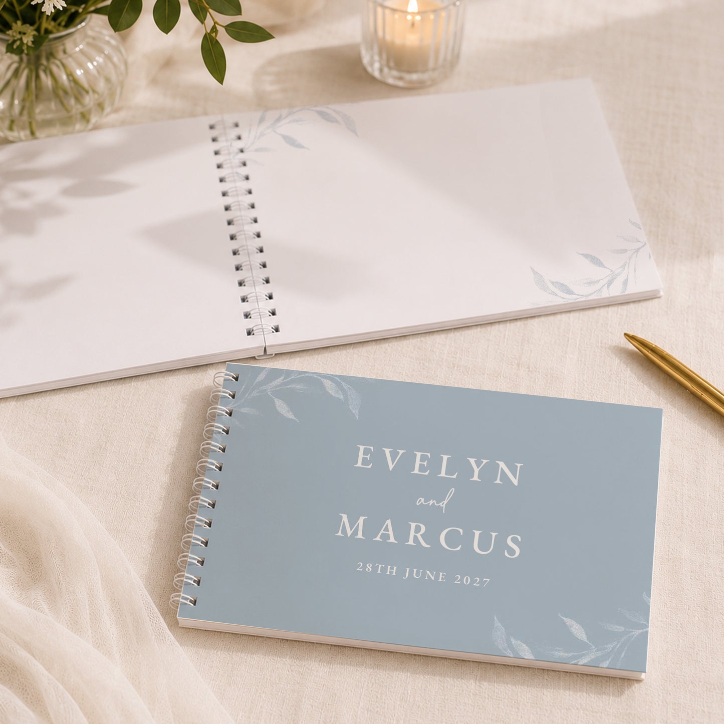 A personalised spiral-bound wedding guest book or agenda in a dusty blue color with white floral accents and the names 'EVELYN and MARCUS' printed on the cover, styled on a beige fabric surface with a gold pen, an open notebook, and a glass candle holder in the background.