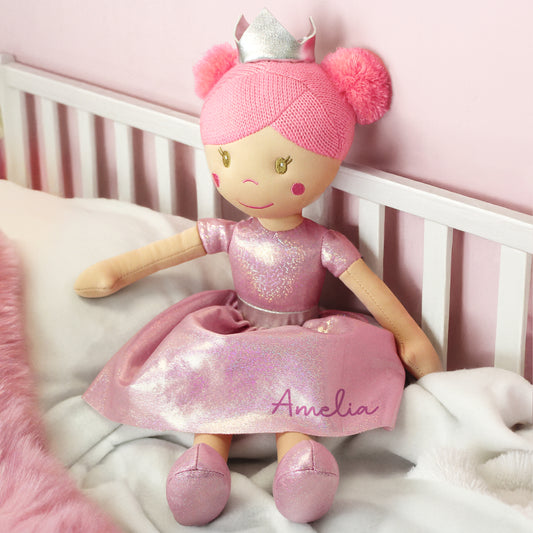 Doll named Amelia in a pink outfit with a crown, sitting on a white crib with a pink background.