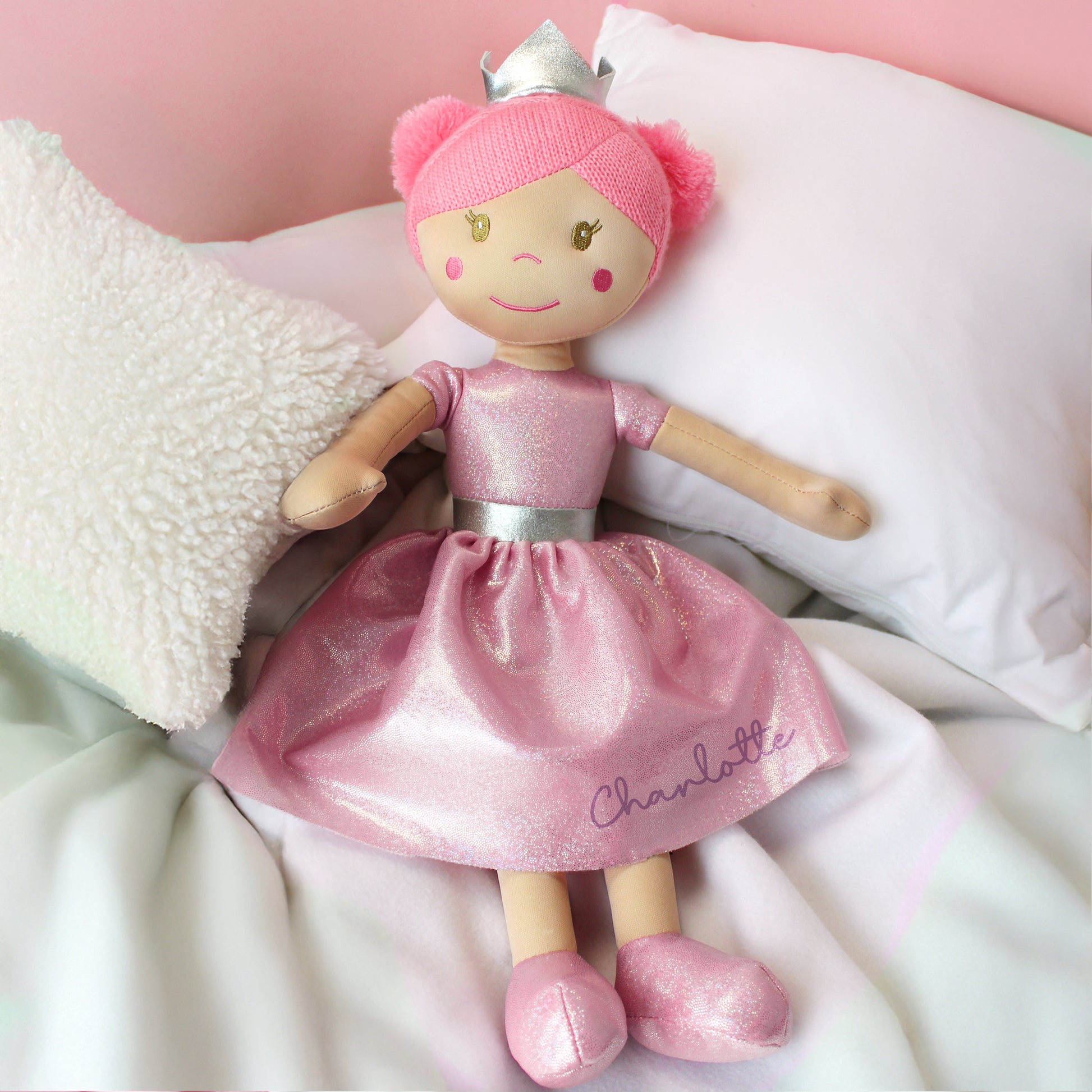 Doll in a pink dress with 'Charlotte' on it, wearing a tiara, on a soft surface with a pink background.