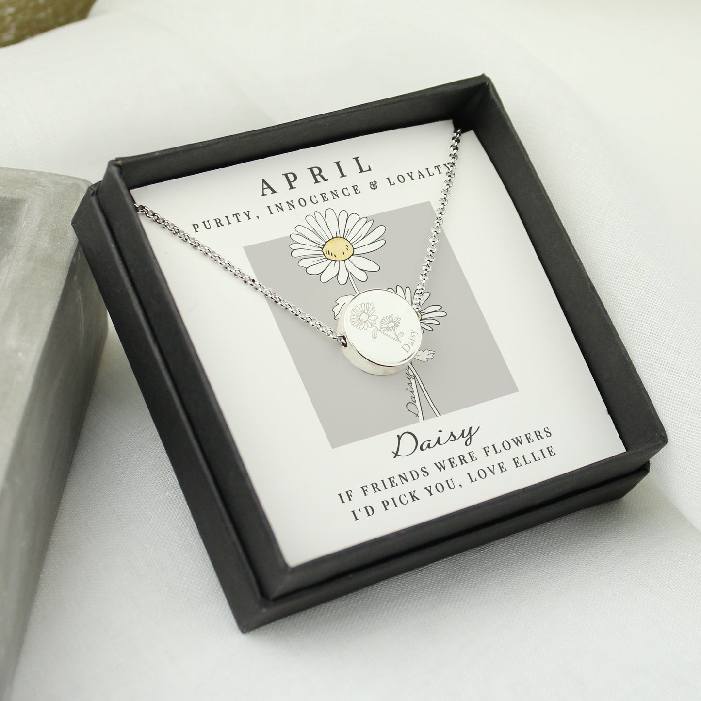 Silver coloured necklace with a daisy pendant in a black box on a white surface