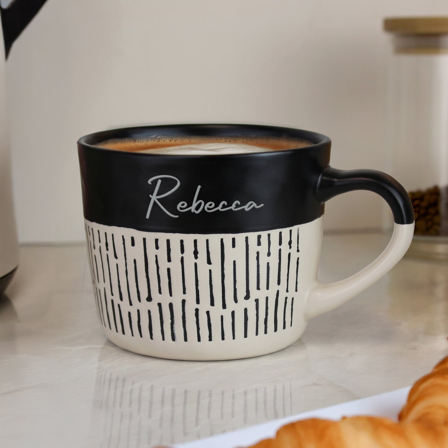 Personalised black and beige mug with 'Rebecca' on a kitchen counter with coffee and pastries.