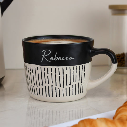 Personalised black and beige mug with 'Rebecca' on a kitchen counter with coffee and pastries.