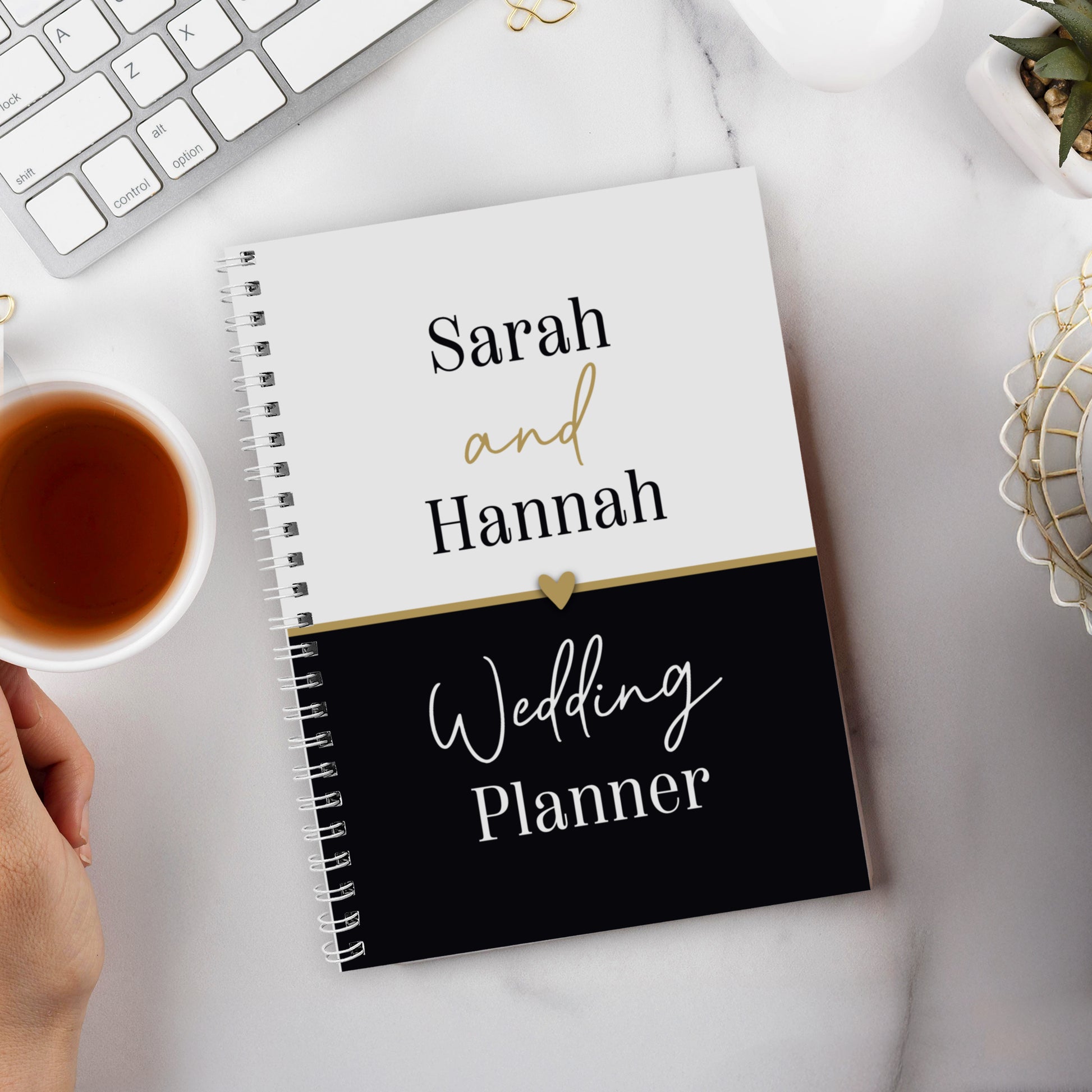 Wedding planner book titled 'Sarah and Hannah Wedding Planner' on a desk with a cup of tea and keyboard.