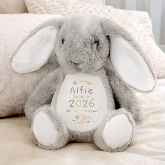 Grey and white soft toy bunny with personalised details - Alfie Born in 2023 8lb 3oz 12.09am among moon and star images. Shown sat on a bed.