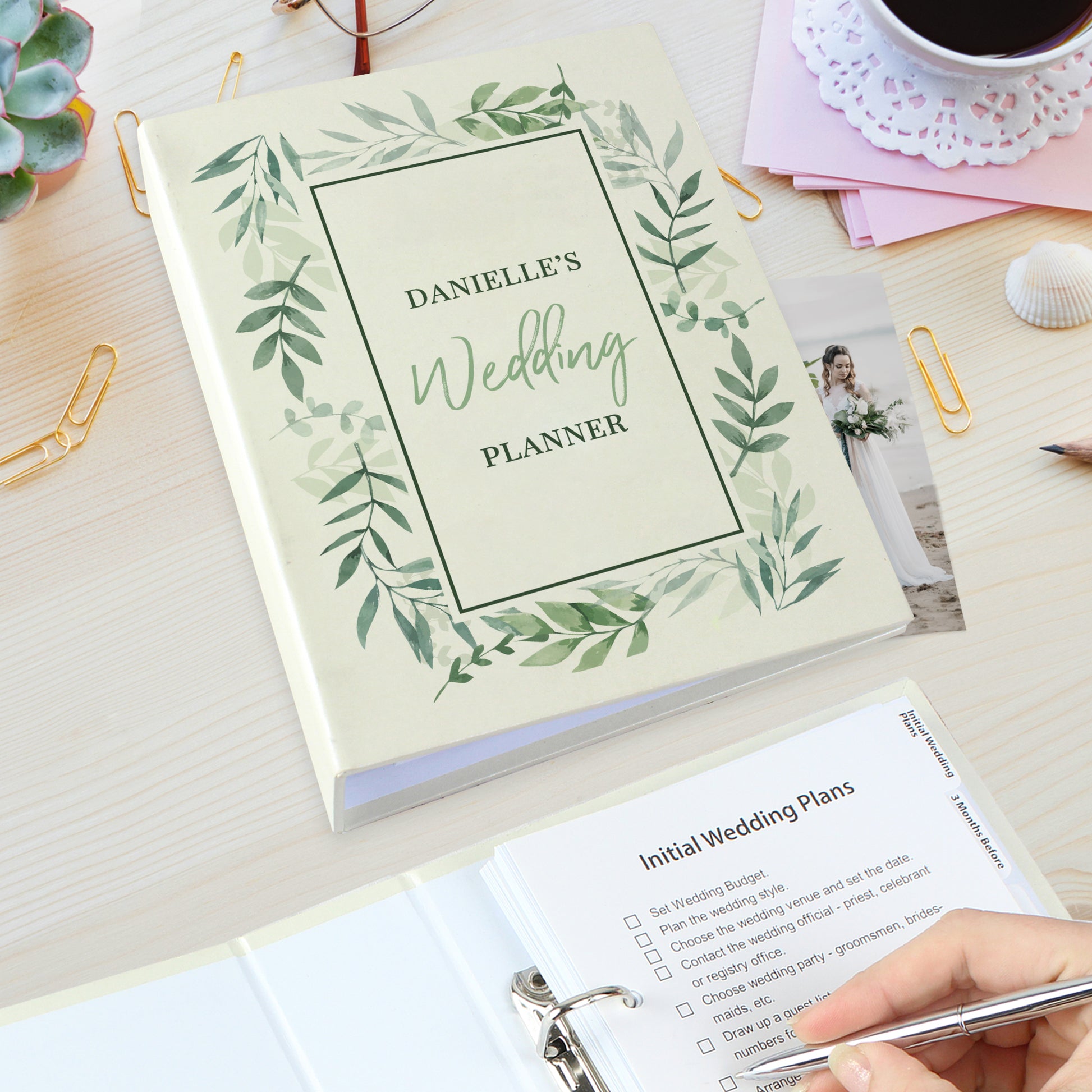 Wedding planner book with green leaf design on a desk with a coffee cup and photos.
