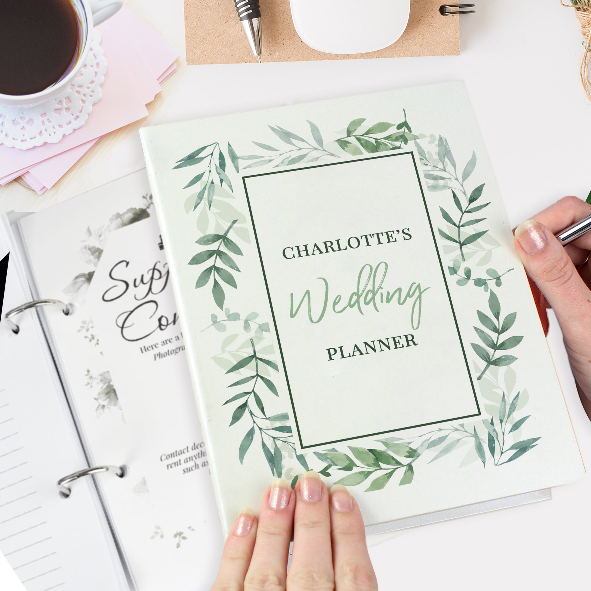 Wedding planner book with green leaf design held by a hand, surrounded by stationery items.