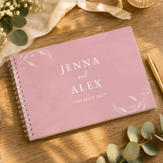 A personalised pink spiral-bound guest book with white text reading 'JENNA and ALEX 7TH JULY 2027' lies on a wooden table surrounded by eucalyptus leaves, a gold pen, and soft decorative props.