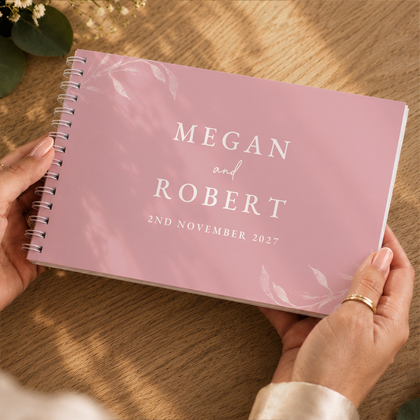 A personalised pink spiral-bound wedding guest book held by two hands over a wooden table. The cover features white text reading 'MEGAN and ROBERT 2ND NOVEMBER 2027' with delicate white leaf illustrations.