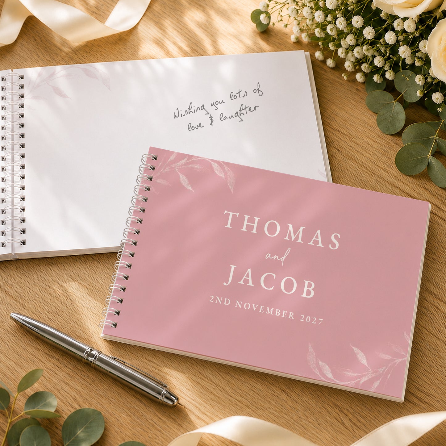 A pink spiral-bound wedding guest book or journal with white text reading 'THOMAS and JACOB 2ND NOVEMBER 2027' is placed on a wooden surface, alongside an open white guest book with handwritten text, a silver pen, and floral decor.