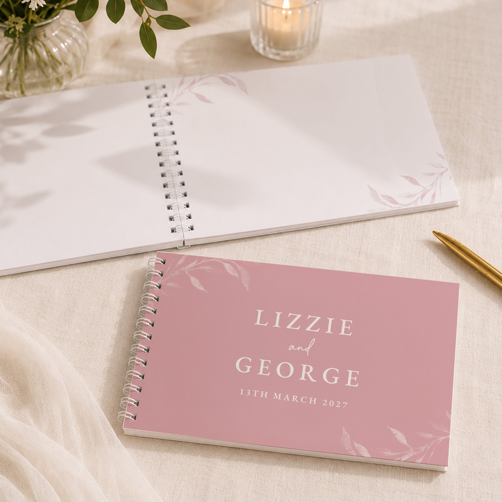 A pink spiral-bound guest book with the names 'LIZZIE and GEORGE' and the date '15TH MARCH 2027' printed on the cover, styled on a light fabric surface with an open white notebook, a candle, and a gold pen nearby.