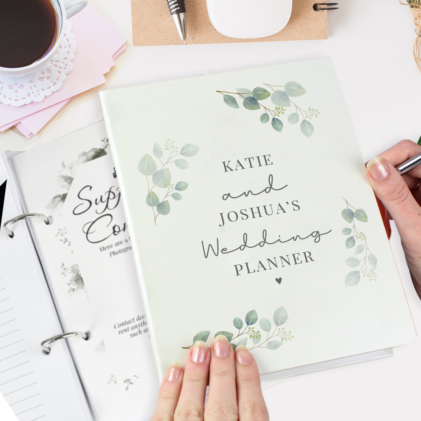 Wedding planner book with floral design held by a hand, surrounded by stationery items.